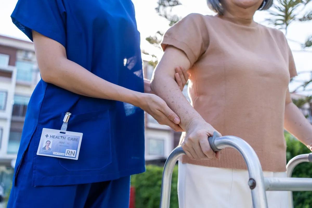 A nurse in blue scrubs is assisting an elderly woman with a walker